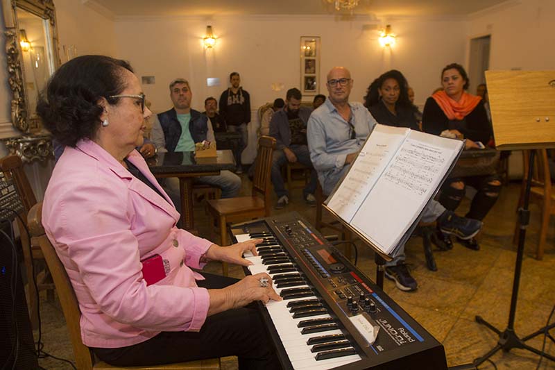 uma mulher sentada tocando piano e outras pessoas em pé ouvindo
