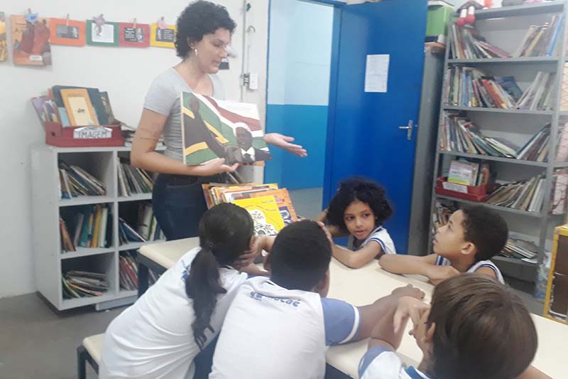 uma mulher com um livro em mãos dentro de uma sala e crianças ao redor